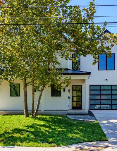 Modern two-story home with a white facade, large windows, and a garage door with glass panels, surrounded by green landscaping.