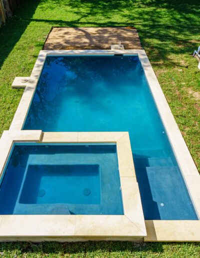 A rectangular swimming pool with an adjacent smaller square pool in a backyard setting, surrounded by grass and a fence.