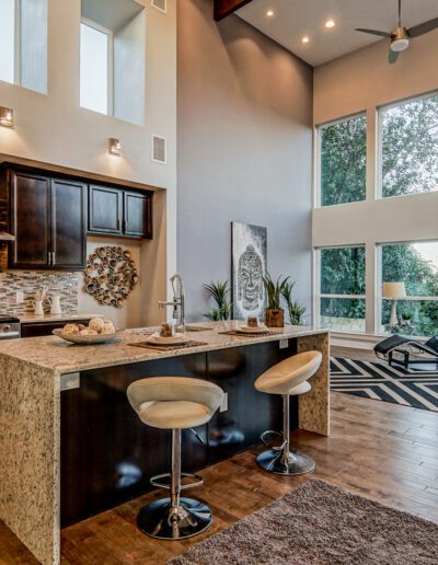 Modern kitchen with bar seating leading into a living area with large windows and a view of trees.