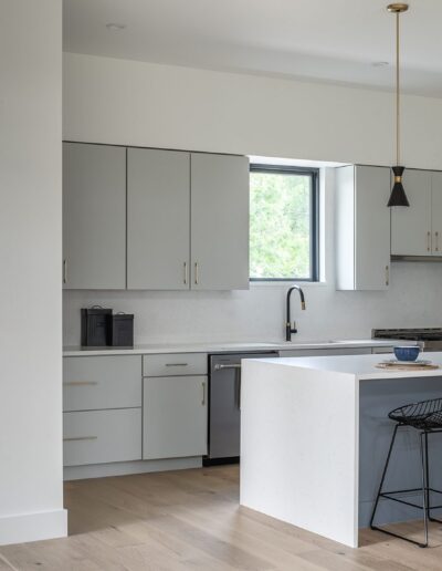 A modern kitchen with light gray cabinets, a central island with a white countertop and black bar stools, pendant lights hanging above, and a window providing natural light.