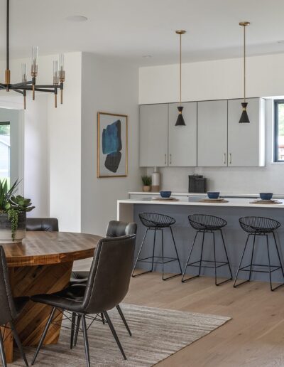 Modern kitchen with a round wooden dining table and six black chairs in the foreground. A kitchen island with four black barstools and pendant lights is in the background, alongside white cabinetry.