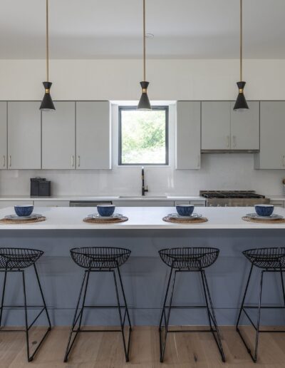 Modern kitchen with a large island featuring four wire-frame stools, white cabinets, pendant lights, a window, and minimalistic decor.