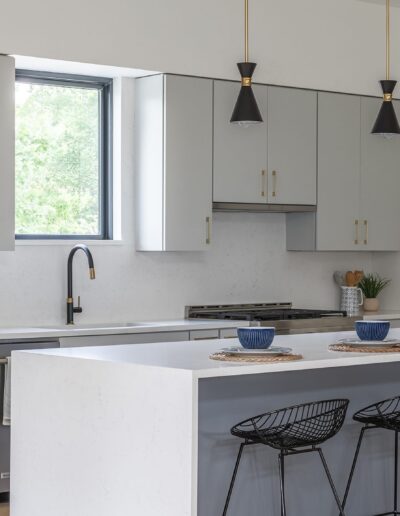 Modern kitchen with white cabinets, a central island with four black bar stools, and a window above the sink. Three pendant lights hang above the island, and blue bowls are set on placemats.