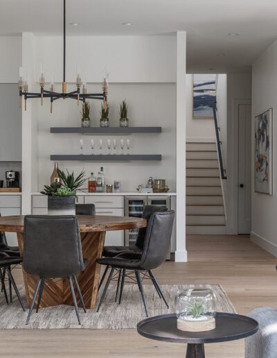 A modern dining area with a round wooden table, black chairs, a chandelier, and kitchen in the background. A staircase is visible in the corner with artwork on the walls.