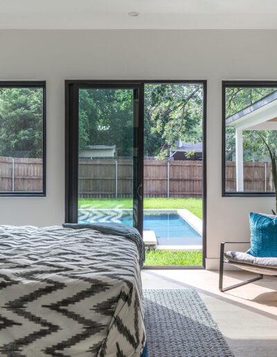 A modern bedroom with a king-size bed, chevron-patterned bedding and pillows, a dresser, an armchair, and a glass door leading to a backyard with a pool. Light streams in through three windows.