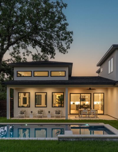 A modern house with large windows is lit up in the evening. The backyard features a rectangular swimming pool, lawn, and patio area with outdoor furniture. A large tree stands near the fenced property.