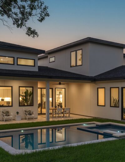 A modern two-story house with large windows, a spacious patio, and an illuminated swimming pool at dusk, surrounded by a well-kept lawn.