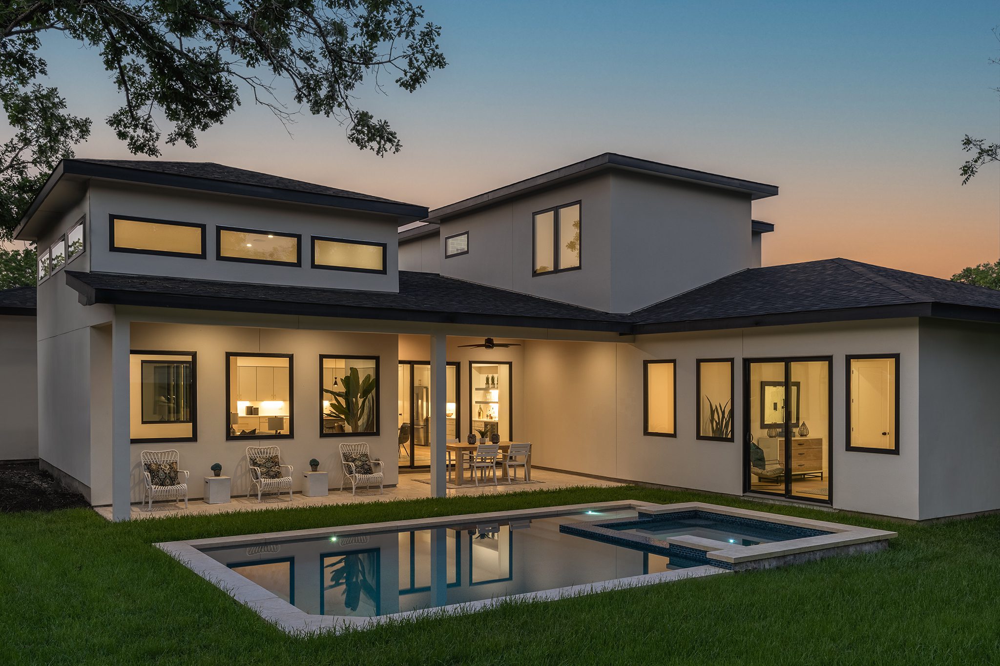 A modern two-story house with large windows, a spacious patio, and an illuminated swimming pool at dusk, surrounded by a well-kept lawn.