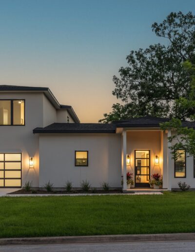 A modern two-story house with illuminated exterior lights at dusk, featuring large windows, a balcony, and an attached garage. A neatly manicured lawn and a tree are in the foreground.