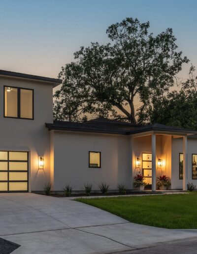 Modern two-story house with a garage, illuminated at dusk. The house features large windows, a balcony, and a manicured lawn with trees.