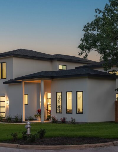 A modern, two-story house with large windows, a flat roof, and well-maintained lawn, illuminated at dusk.