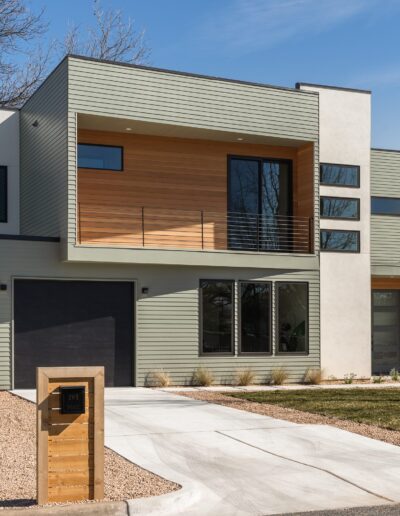 Modern two-story house with a mix of wood and metal siding, large windows, a balcony, and a single-car garage. The front yard features a gravel path, a mailbox, and a driveway.