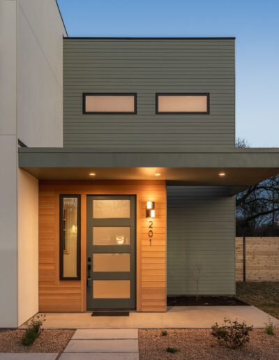 A modern house entrance with a lit wall lamp above the door number 201. The door is flanked by wooden panels and a small porch area is visible in the foreground.