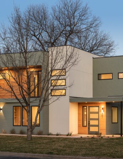 Modern two-story house with large windows, a mix of light beige and gray exterior, a wooden accent wall on the upper level, a garage, and illuminated entrances. There's a leafless tree in the front yard.