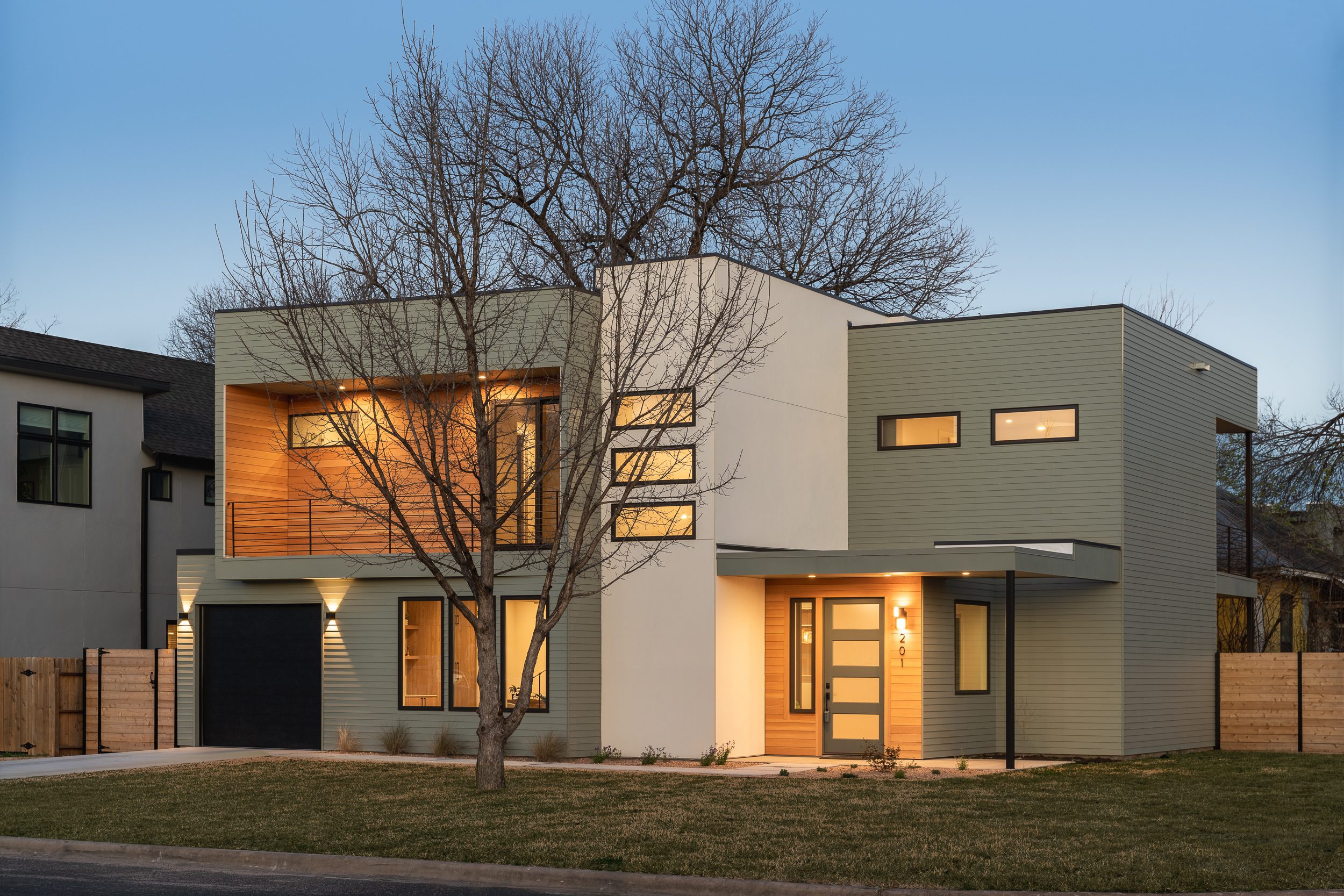 Modern two-story house with large windows, a mix of light beige and gray exterior, a wooden accent wall on the upper level, a garage, and illuminated entrances. There's a leafless tree in the front yard.