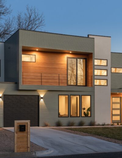 Modern two-story house with large windows and wood accents, illuminated at dusk. The house features a front lawn, driveway, and a wooden fence with a mailbox.