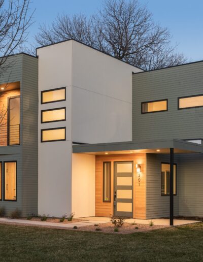 Modern two-story house with clean lines, featuring a mix of light gray and white exterior, large windows, and a covered front entryway, set against a backdrop of leafless trees and a clear evening sky.