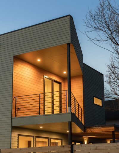 Modern two-story house with a balcony and railing, illuminated by warm lights, against a twilight sky with bare tree branches in the background.