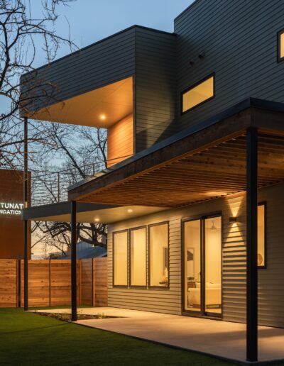 Modern two-story house with large windows and a covered patio, illuminated during twilight. A wooden privacy fence and bare tree branches are visible in the background.