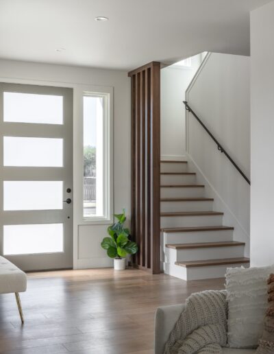 A modern entryway with a white door featuring frosted glass panels, a small plant in a white pot, a wooden staircase with a black handrail, and a portion of a white sofa on the bottom right.