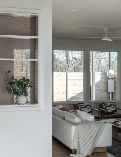 A modern living room with a white couch, a coffee table, and a beige throw blanket. A potted plant sits on a shelf in the foreground. Large windows and a wooden fence are visible in the background.