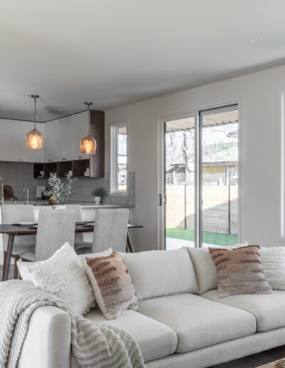 Modern open-plan living area with a white sofa, light wood floors, and a kitchen featuring white cabinetry, a large island with barstools, pendant lights, and a dining table. Large windows illuminate the space.