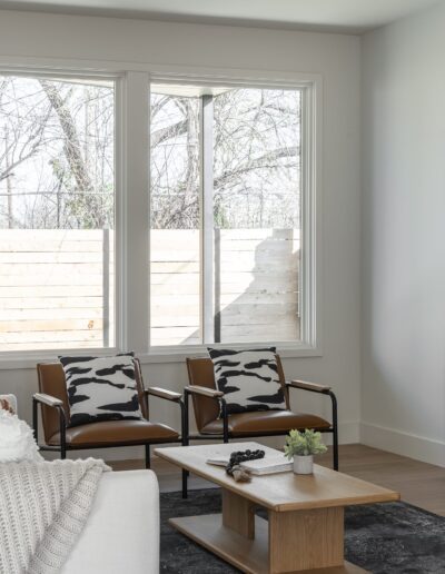 A modern living room with a white sofa, two brown armchairs, a wooden coffee table, and a TV console. Large windows on the left side let in natural light. Minimalistic decor with neutral tones.