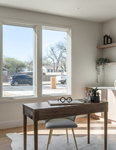 A modern home office with a wooden desk, chair, and storage cabinets. A large window is behind the desk, and a potted plant and floor lamp are to the left.