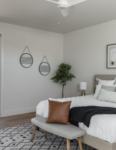 A modern bedroom with a large bed, white bedding, a brown pillow, two side tables with lamps, a bench, a geometric-patterned rug, a potted plant, two wall mirrors, and framed artwork. Large glass doors to the left.