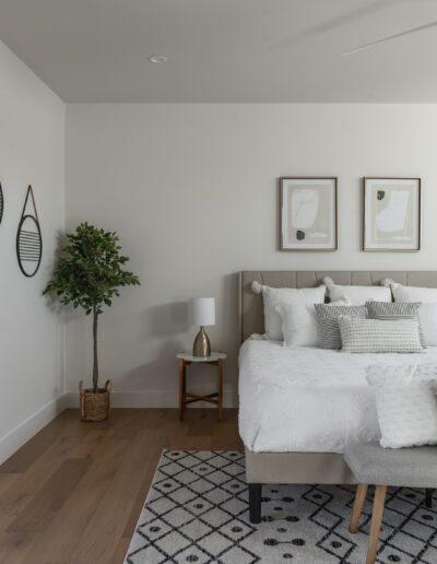 A modern bedroom with a bed, nightstands, decorative pillows, and a bench. The room features a potted plant, wall art, a patterned rug, a sliding glass door, and natural light from a window.