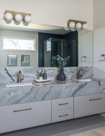 A modern bathroom features a marble double vanity with silver fixtures, a large mirror with globe lights, and decorative plants. The shower has dark tiles and a small window is above the vanity.